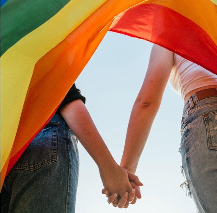 Two individuals holding hands under a rainbow Pride flag, symbolizing LGBTQ+ inclusion, diversity, equality, and support for human rights.