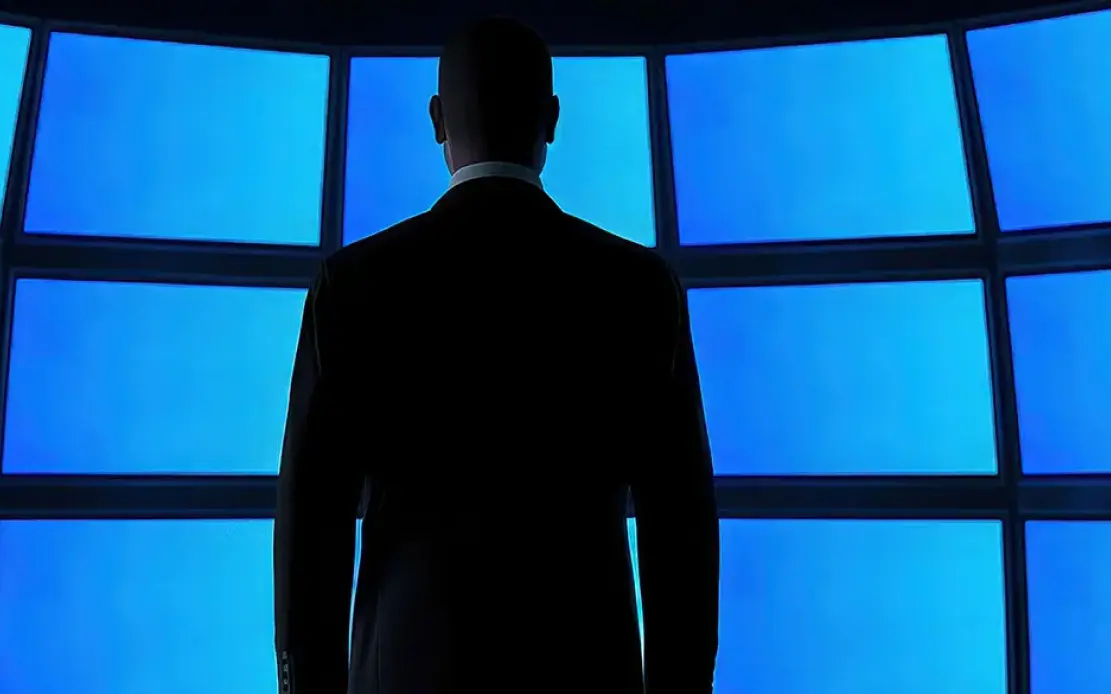 A person in a suit stands before large blue screens, viewed from behind.
