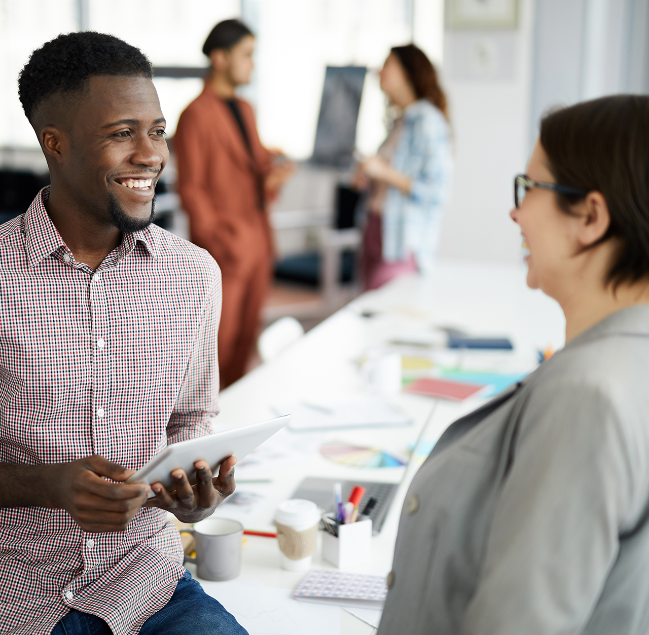 Zwei Studenten stehen in einem modernen Büro neben einem Schreibtisch und unterhalten sich. Eine Person hält ein Tablet in der Hand, während auf dem Schreibtisch Papiere, Diagramme und Büromaterial zu sehen sind. Im Hintergrund interagieren andere Personen neben einer Weißwandtafel und schaffen so eine kollaborative Arbeitsumgebung.